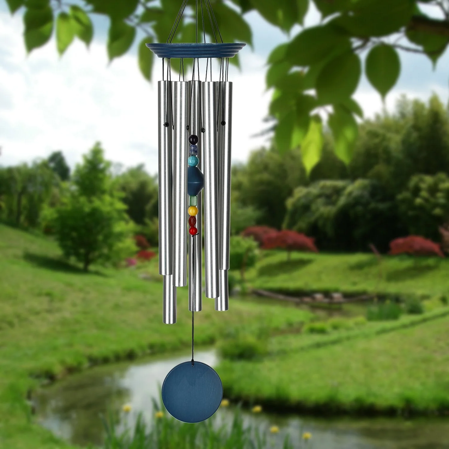 Woodstock Percussion 7‑Stone Bronze Chakra Wind Chime with engravable wooden wind sail WITH seven SILVER aluminum tubes hang from BLUE WASH finish ash‑wood top; the faceted clapper aligns seven chakra stones vertically, with a round wooden sail below that can be personalized with up to 50 characters