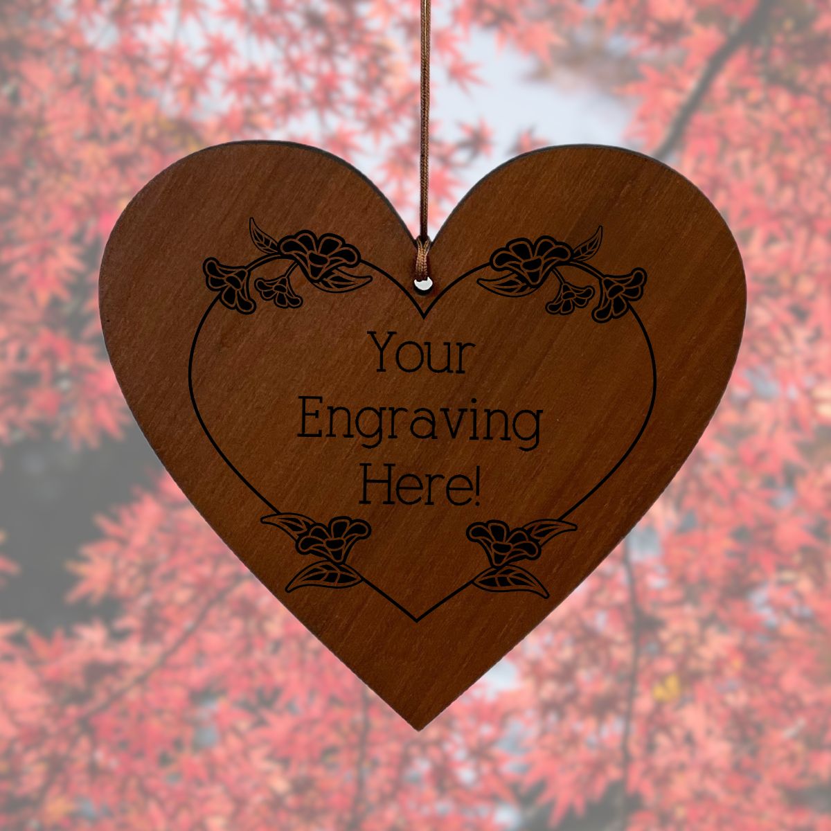 Wooden heart-shaped engraving plaque with floral designs and 'Your Engraving Here!' text, against a blurred natural background.