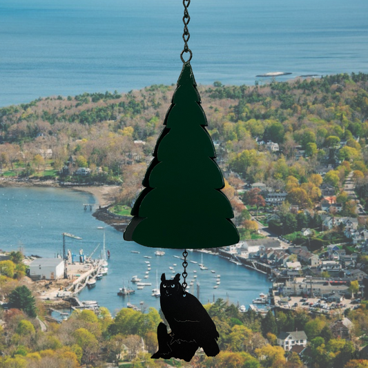 North Country MOUNT BATTIE Buoy Bell – an 8-inch powder‑coated recycled steel bell in a triangular shape WITH CUT OUTS ON THE EDGE GIVING THE BELL A PINE TREE LOOK, hanging from jack chain with customizable windcatcher sailing below; finished in GLOSSY GREEN that weathers into rich rust tones DISPLAYED IN FRONT OF A LANDSCAPE BACKGROUND