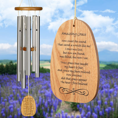 Woodstock Reflections Amazing Grace wind chime with silver tubes and an inscription on the wind sail that reads "Amazing Grace, How sweet the sound
That saved a wretch like me!
I once was lost, but now am found;
was blind, but now I see.
'Twas grace that taught my heart to fear,
And grace my fears relieved;
How precious did that grace appear
The hour I first believed." on a blurred background of a field of blue flowers and blue sky